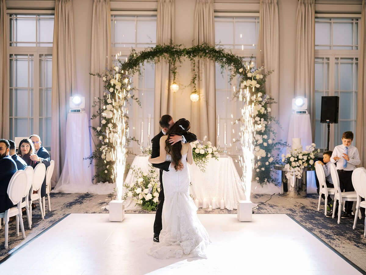 A couple in wedding attire shares their first dance in an elegant venue decorated with flowers and a sparkling backdrop, surrounded by seated guests. This classic Calgary wedding at the Fairmont Palliser exudes timeless charm and sophistication.