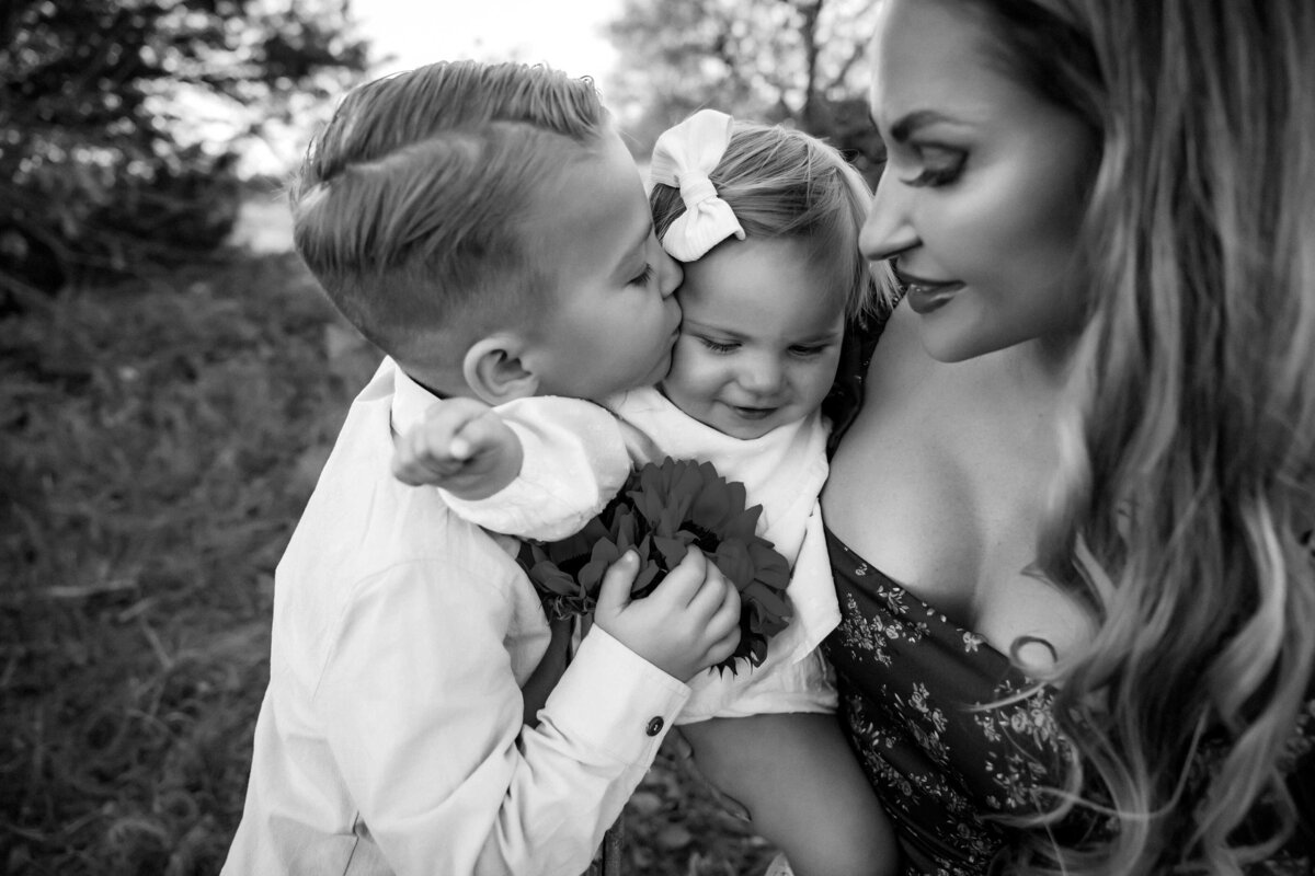 black and white photo of a mother, her baby girl and her son. Baby girl is wearing a white dress and a bow.