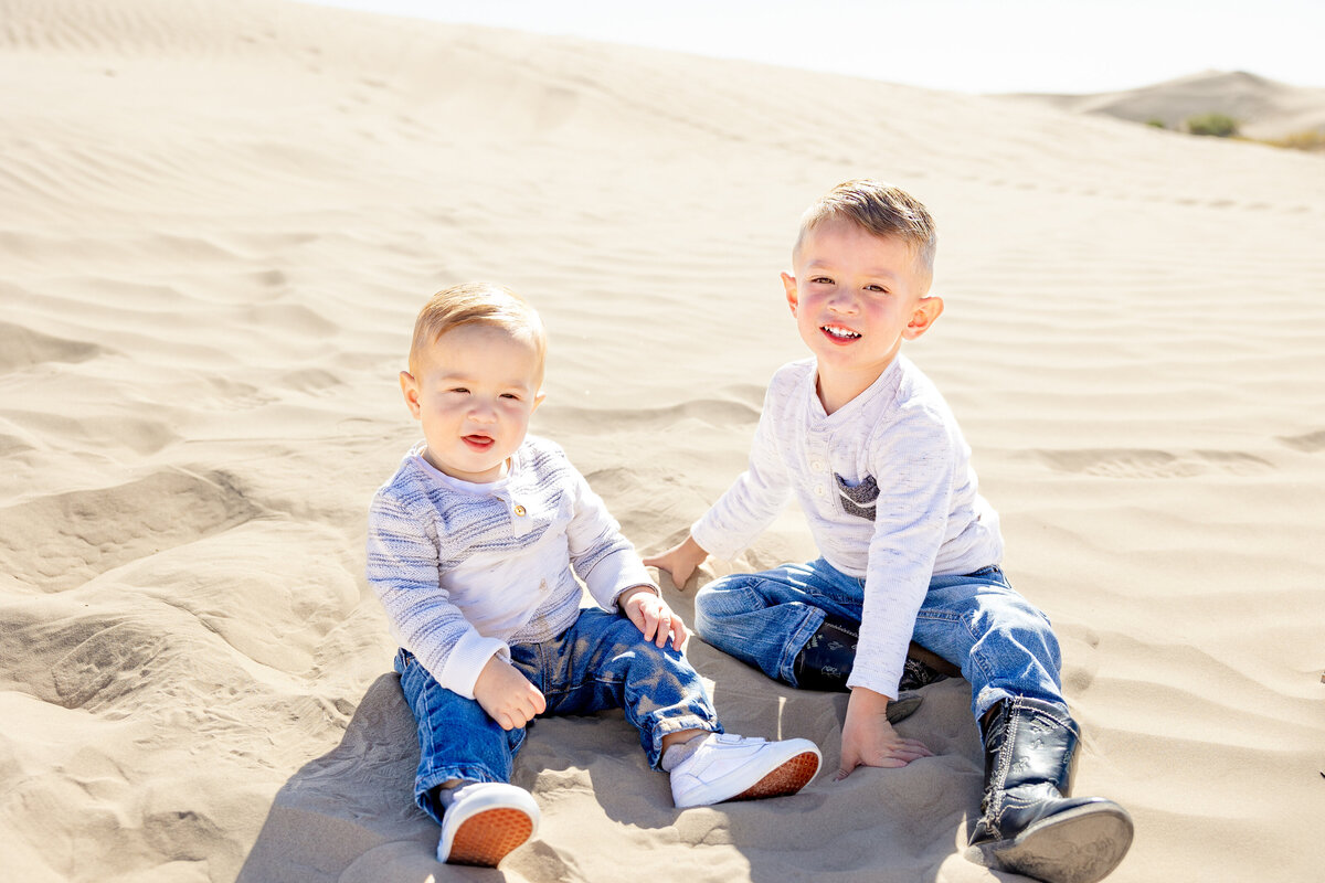 Two young brothers sit on the warm desert sand, laughing and holding hands. Their matching sweaters and jeans contrast with the soft golden tones of the dunes stretching behind them.