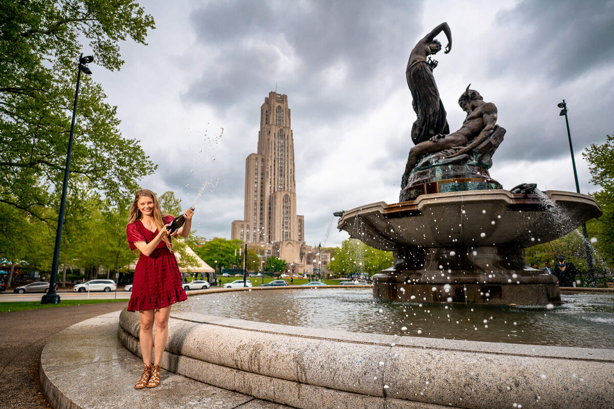 college graduation photography of a woman popping champagne by a water fountain at the university of Pittsburgh. Captured by graduation photographer Michael Fricke Photography.