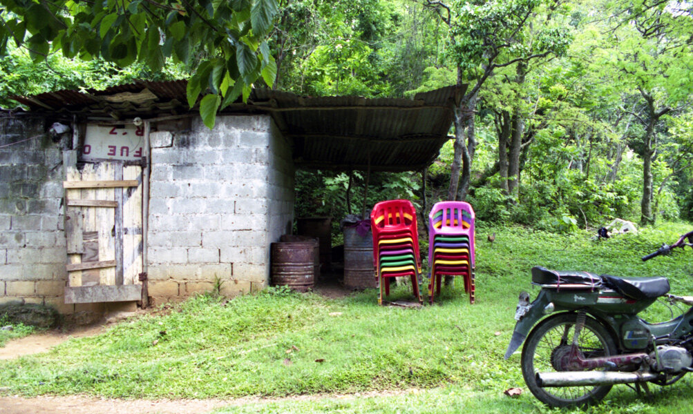 Bright plastic chairs are stacked next to makeshift dilapidated building with forest in background