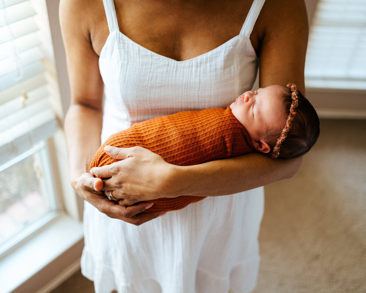 Photograph of a woman in a white dress holding a newborn baby girl with her eyes closed, covered with an orange blanket and holding a didema on her head