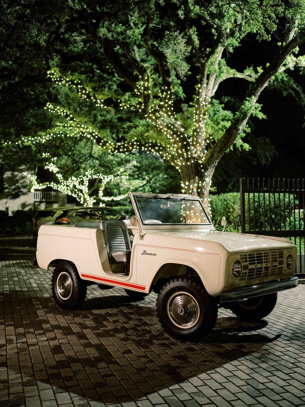 A classic vintage Bronco parked elegantly under softly lit trees at night, showcasing its retro charm, as lights twinkle, creating a romantic ambiance in a serene outdoor setting.