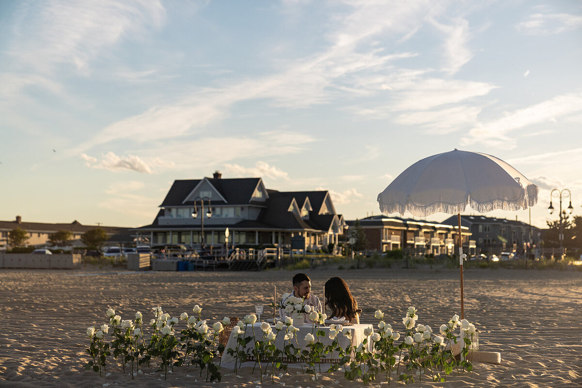 A romantic beachside dinner setup with white roses, a round table, and a fringe umbrella, styled by Yours Truly, a New Jersey wedding planner and designer.