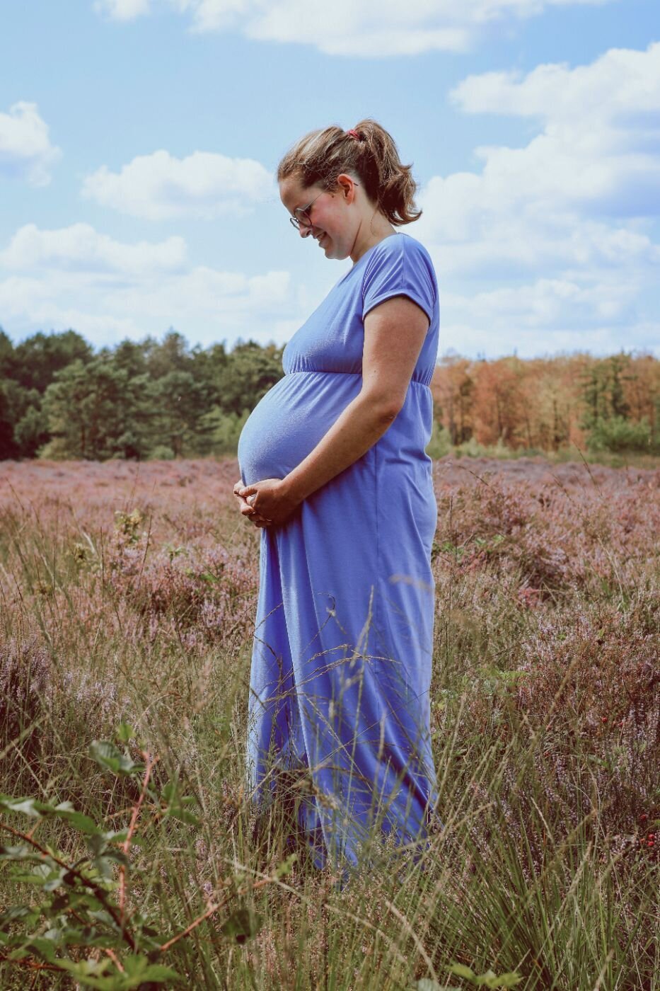 Een zwangerschapsportret van een vrouw in een blauwe jurk, staand in een heidelandschap