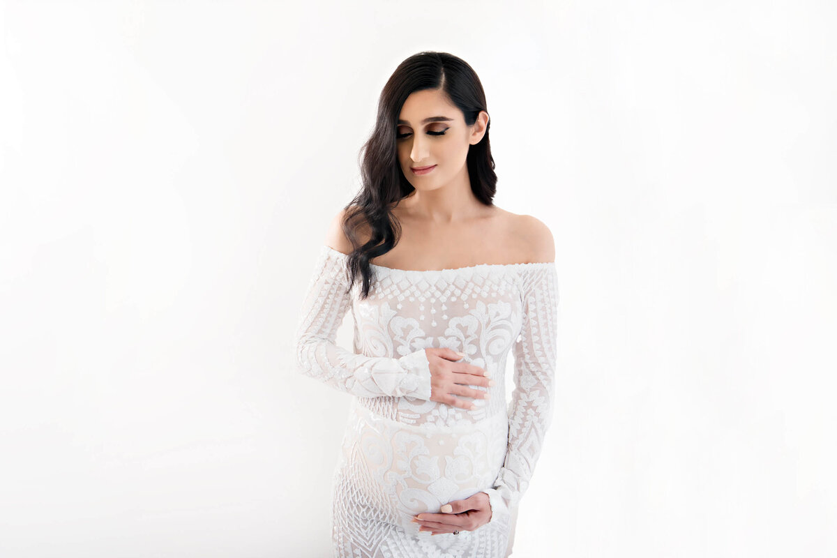 Brown-haired pregnant woman cradling her belly in a white maternity dress on a white background for her Burlington, ON studio maternity photography session