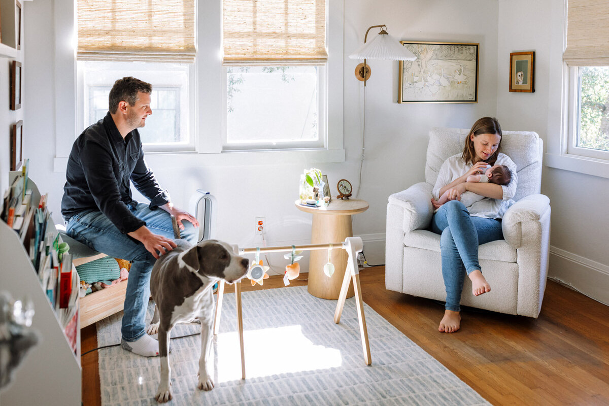 mom sitting in chair feeding bottle to newborn while dad sits in another chair watching and petting the dog