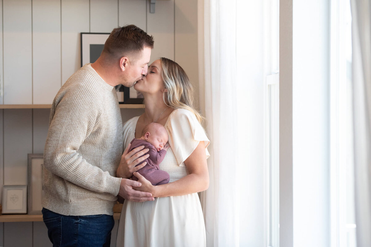 Mom and dad standing in a window kissing while holding their sleeping newborn baby