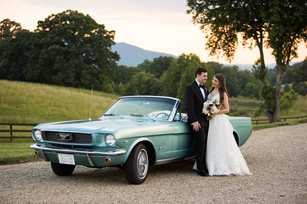 wedding couple stands next to a classic car as the sun sets behind the montains