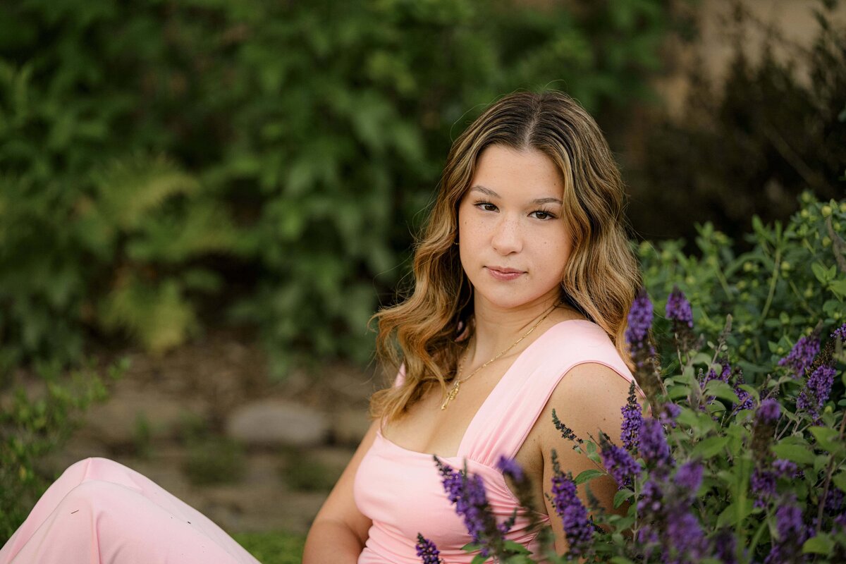 A cuthbertson high school senior dressed in a pink dresses sitting in a flower bed surrounded by purple blooms in her senior session.