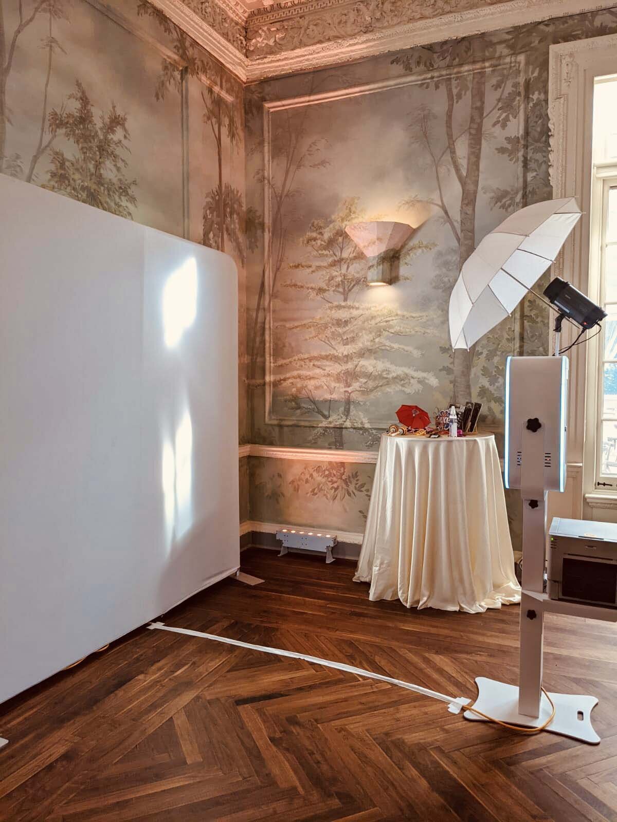 White photo booth with umbrella lighting and white backdrop in a vintage-style ballroom with mural walls.