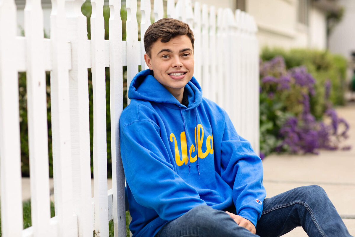 A young person with short brown hair is sitting on the ground against a white picket fence, smiling. They are wearing a blue hoodie with UCLA printed in yellow and jeans. Purple flowers and green bushes are in the background.