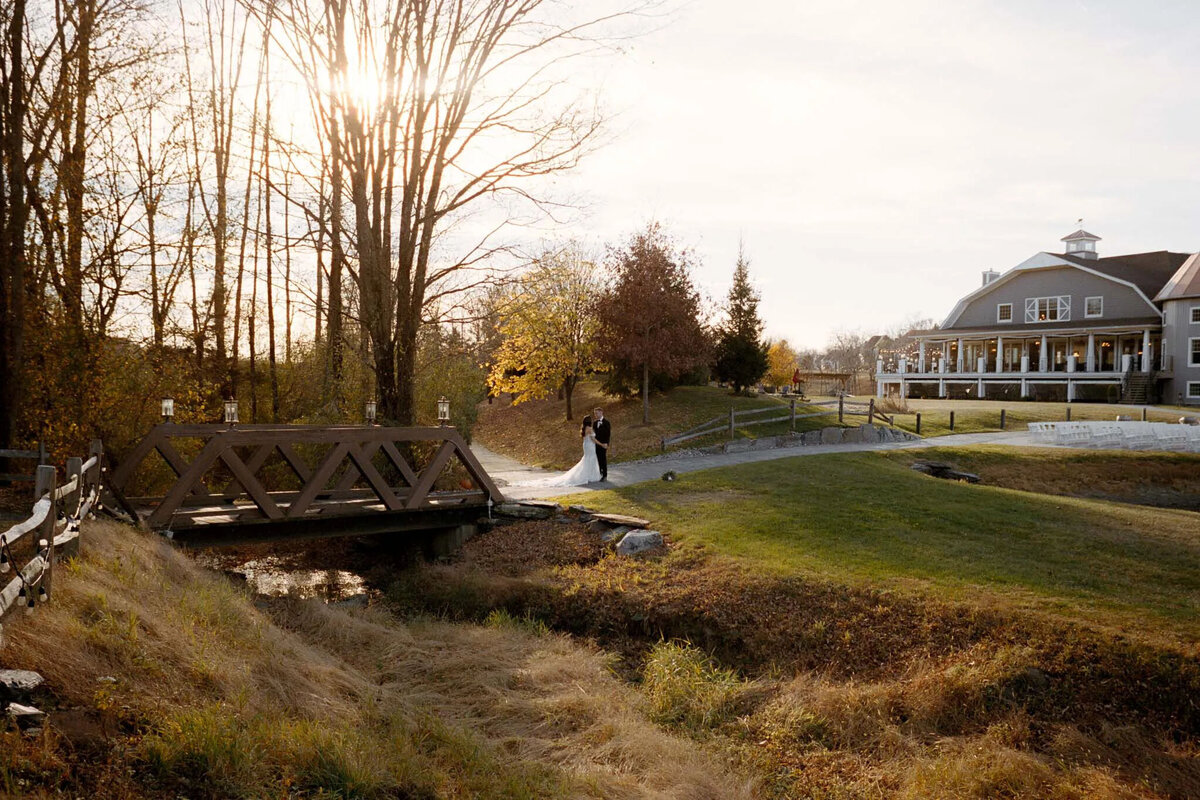 A bride and groom stand together near a wooden bridge, surrounded by autumn trees and sunlit grass, as a nj wedding photographer captures the moment with a large farmhouse in the background.