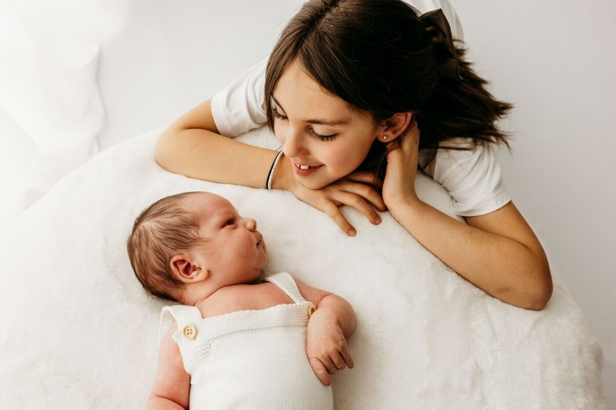 an older sister lovingly gazing at her newborn sibling lying on a soft white blanket. A tender sibling moment captured by Ruaa Hamdan