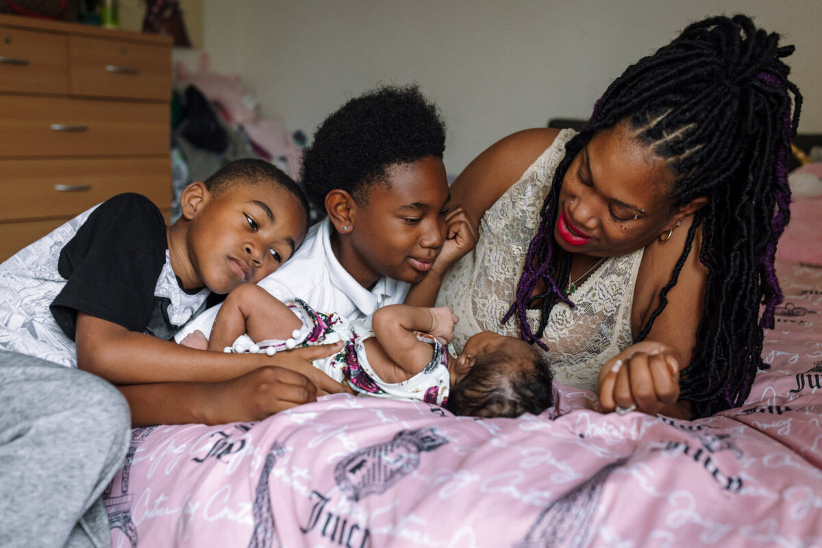 mom and two older brothers laying on pink comforter looking at newborn sister