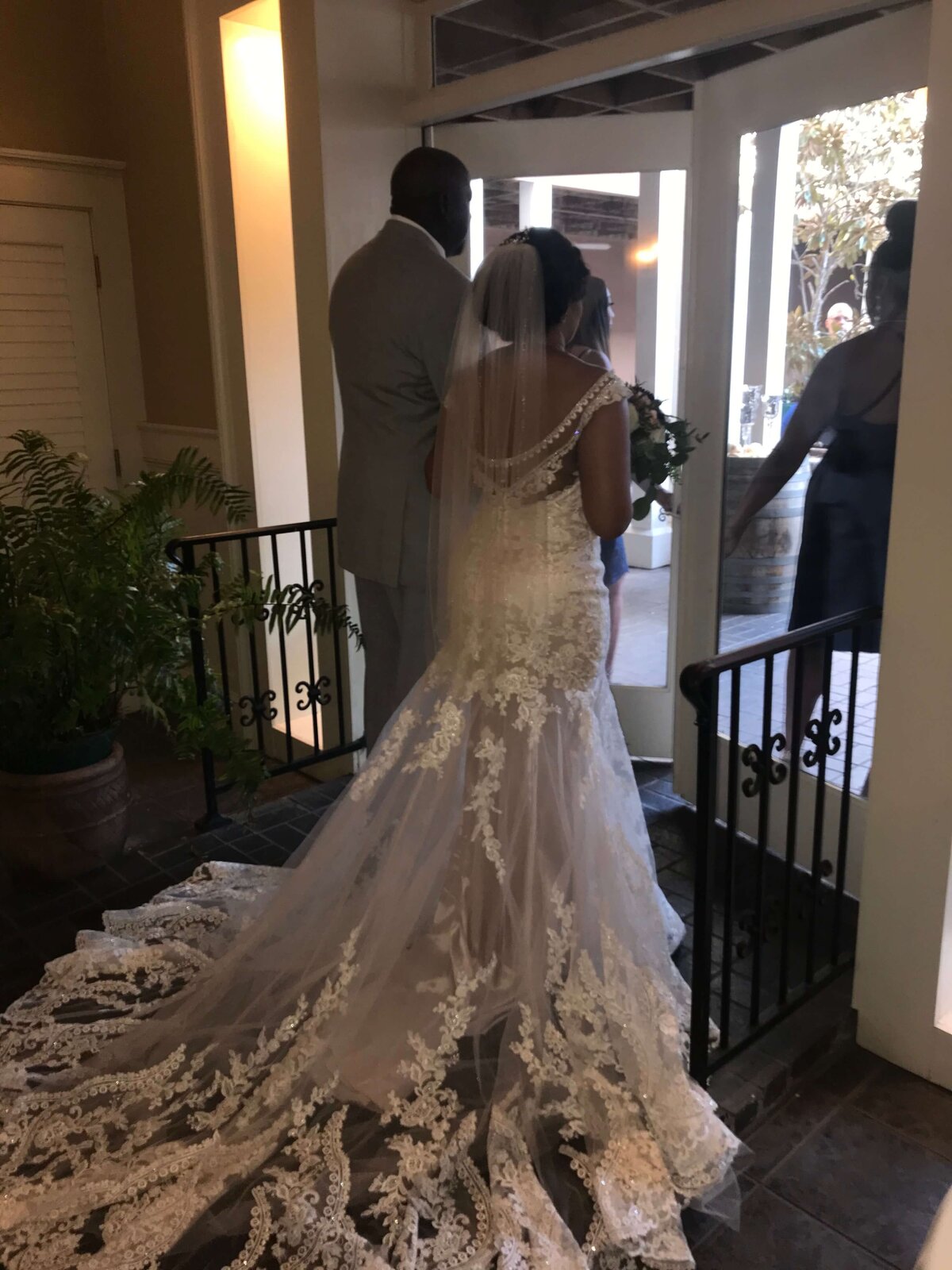 A bride in a white, lace wedding gown with a long train and veil stands beside a man in a suit, preparing to walk through a doorway to an outdoor area where people are waiting.