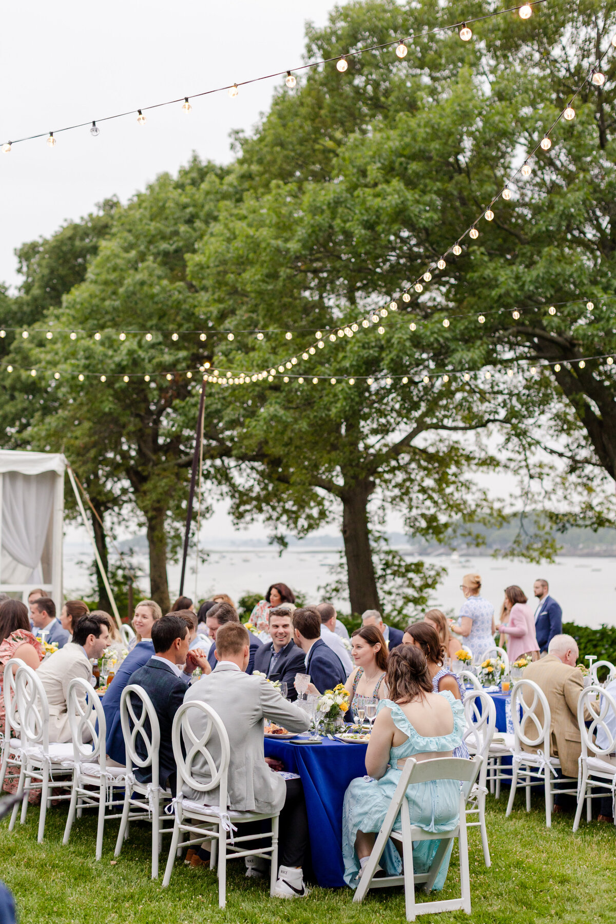 view of the outdoor  rehearsal dinner under string lights and guests laughing at long tables on the lawn. 