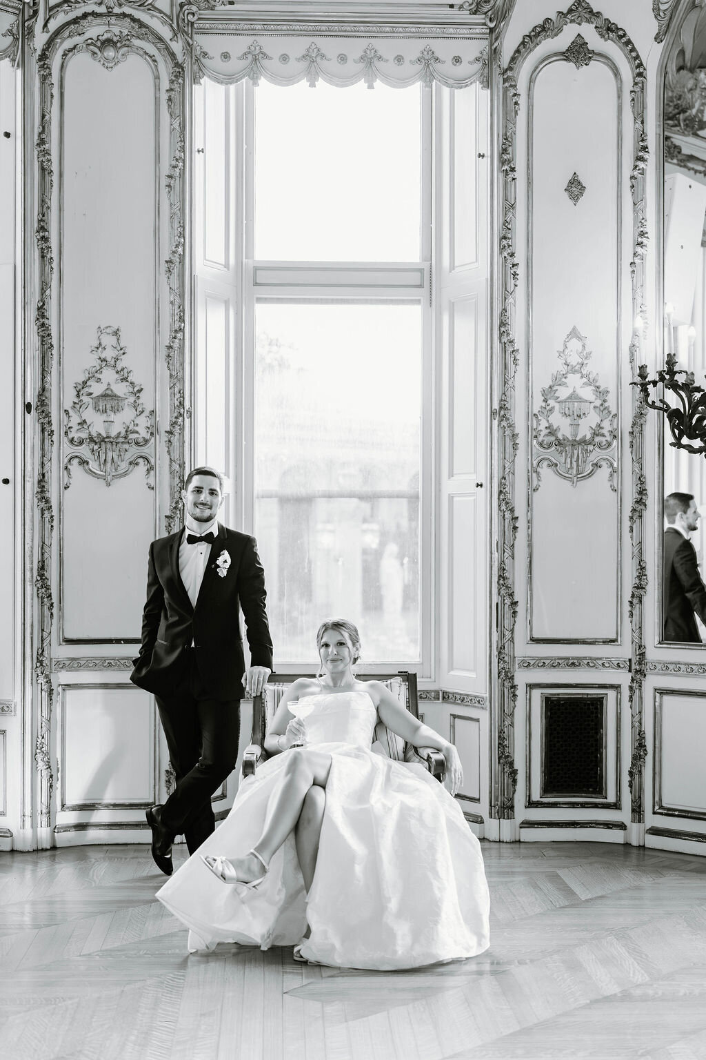 Rhode Island Wedding Photographers | A formally dressed couple poses in an elegant, ornate room. The man stands in a suit, leaning on a chair, while the woman sits in a wedding gown.