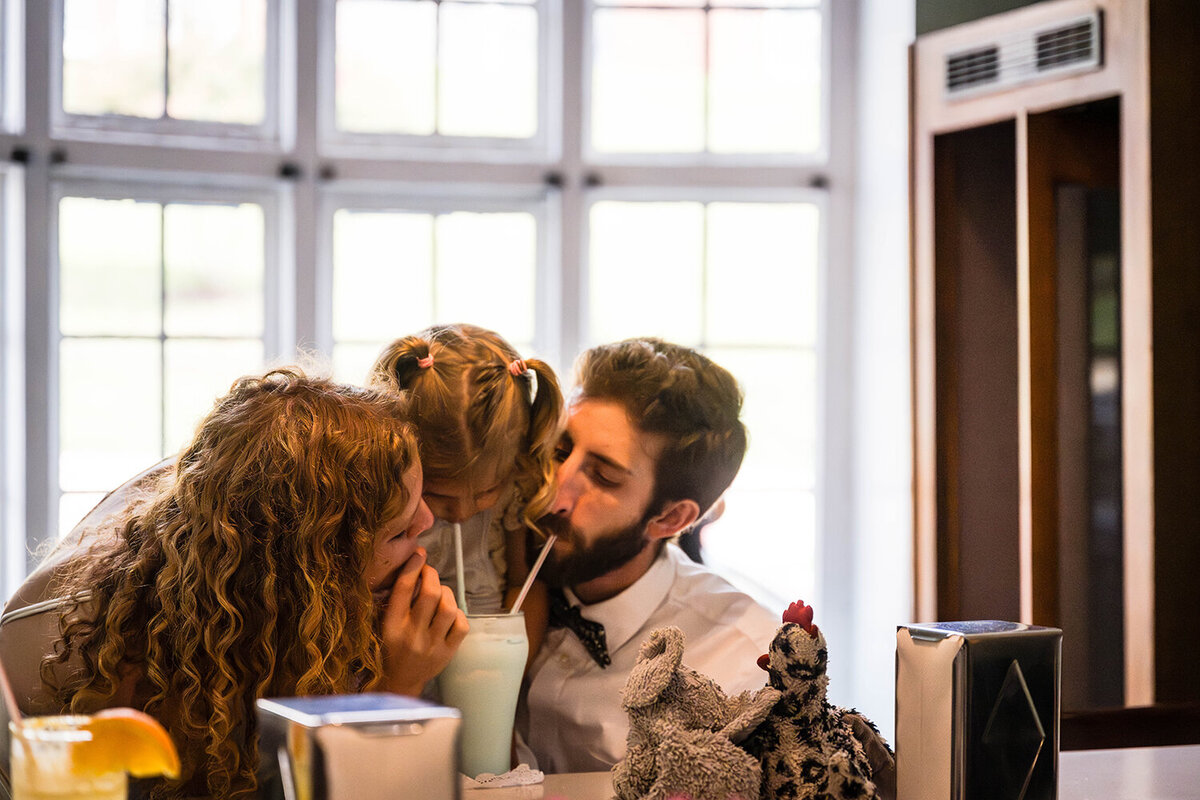 A mother, a daughter, and a father drink from a milkshake together at the bar of Pop’s Ice Cream and Soda Bar in Grandin Village in Roanoke, Virginia following their adventure at Fishburn Park for their elopement day.