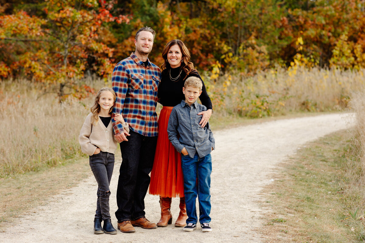A family of four stands on a dirt path in an autumn landscape, captured beautifully by Amber Bauer Photography. The parents are in the middle, with two children on either side. The vibrant trees showcase fall colors. They are all smiling, donning casual fall attire befitting a Brainerd Lakes Area photographer's portfolio of family portraits.