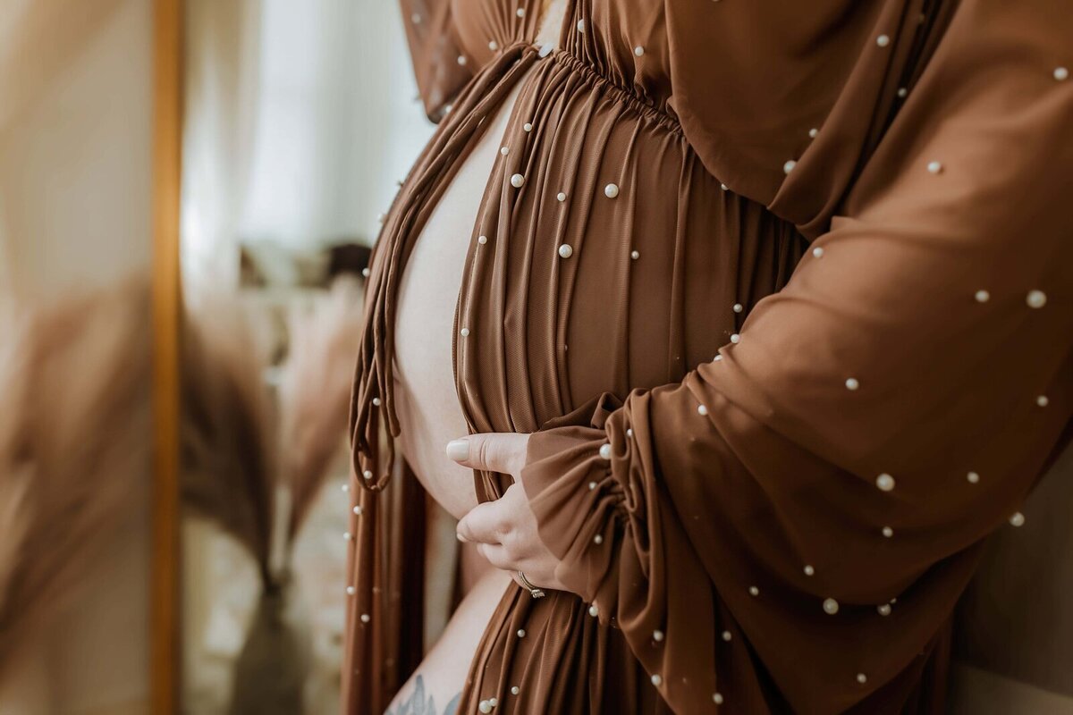 A person wearing a brown, pearl-embellished dress holds their pregnant belly with one hand by a mirror.