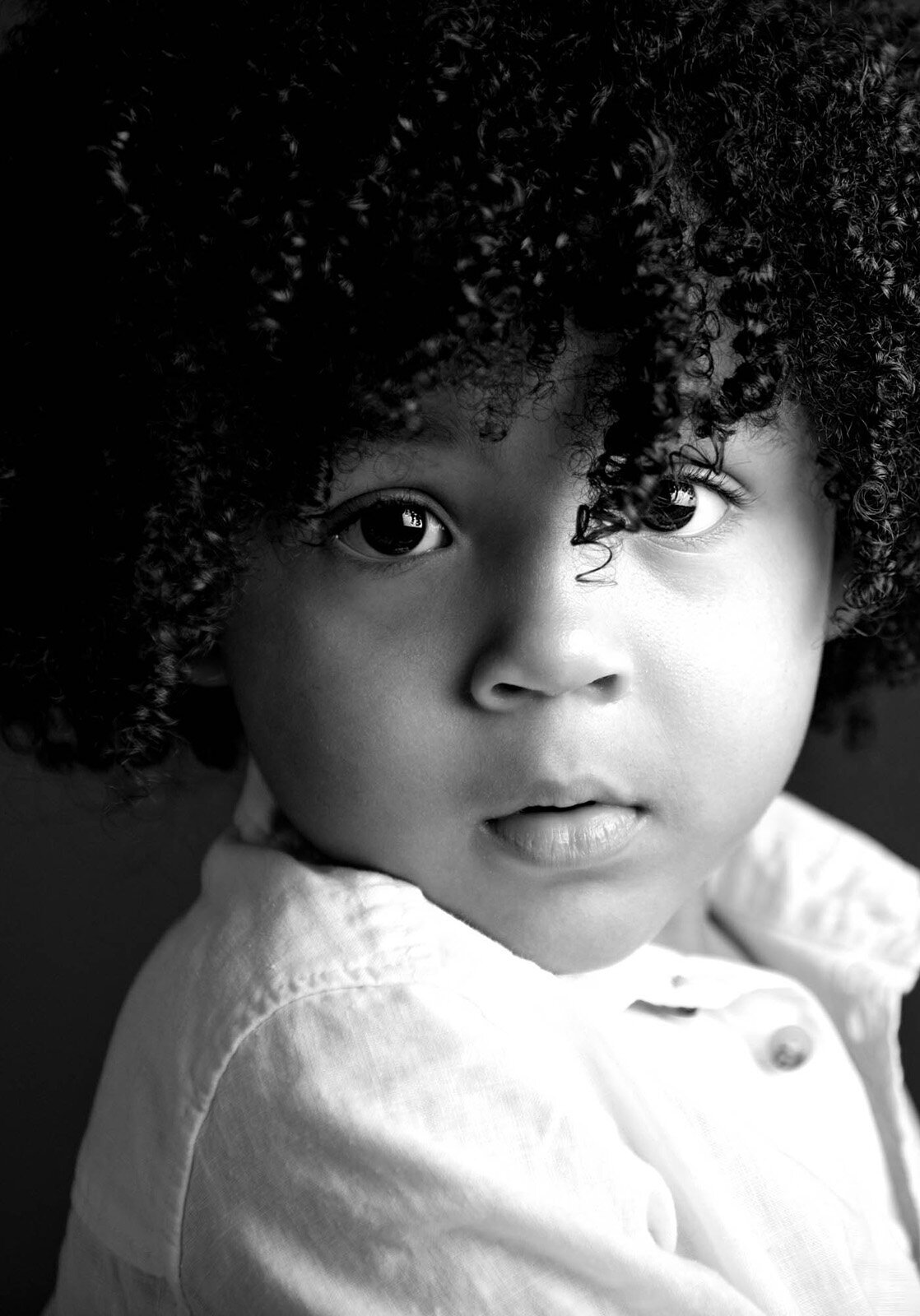 A black and white photo of a young child with curly hair, smiling and looking playfully at the camera.
