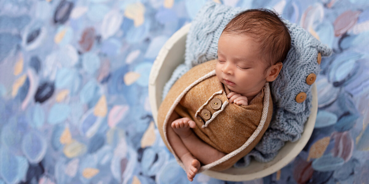 Newborn baby boy wrapped in warm yellow fabric and posed in a white bowl with a blue knit layer, set against a blue textured painted backdrop.