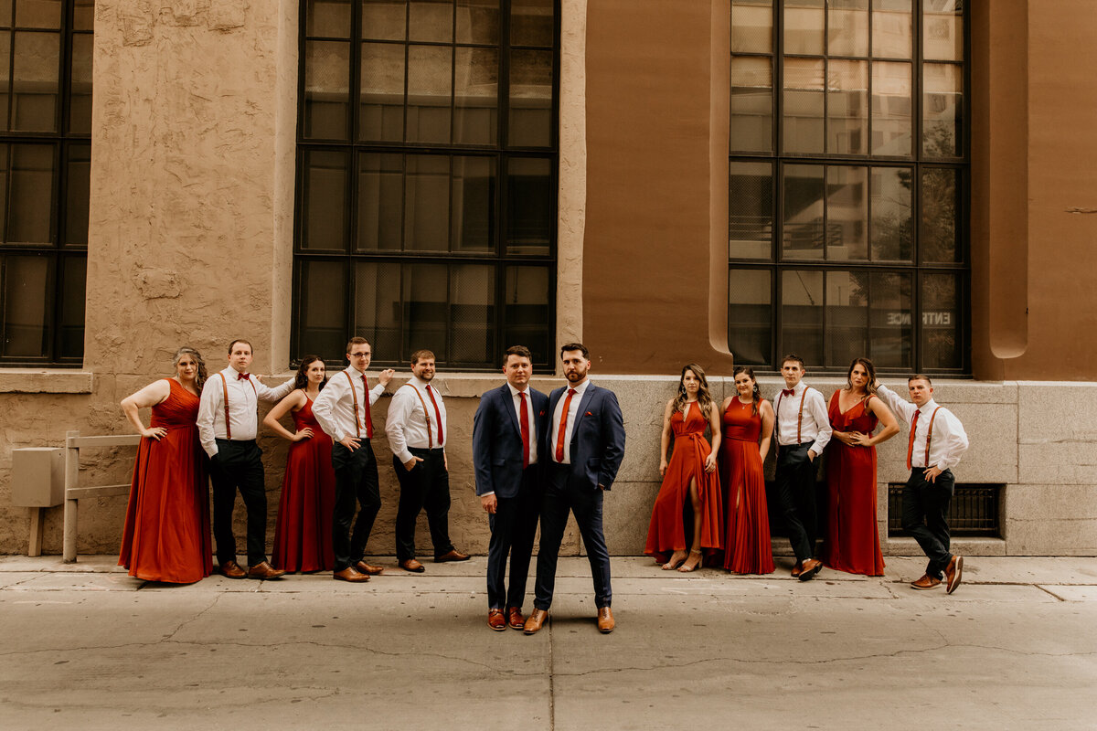 two grooms standing in front of a large window of a brick Albuquerque building with their wedding party