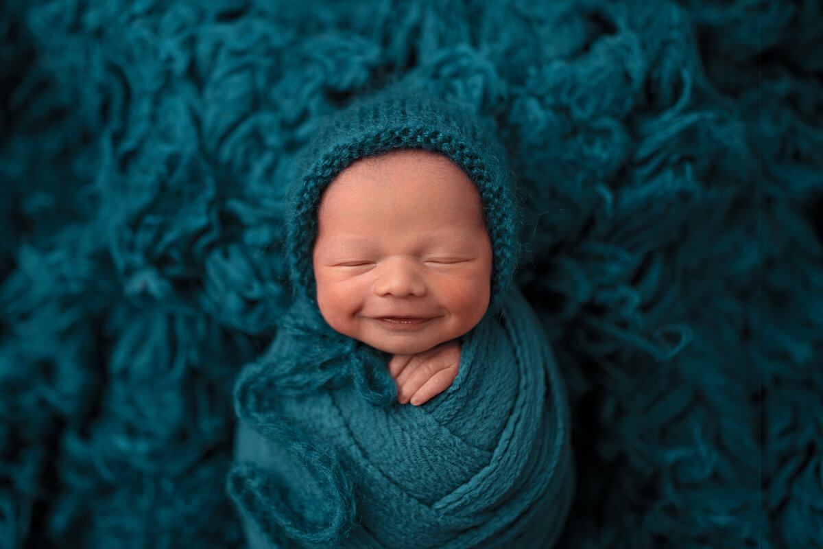 Newborn baby boy smiling while wrapped in teal on fluffy textured backdrop for studio session.