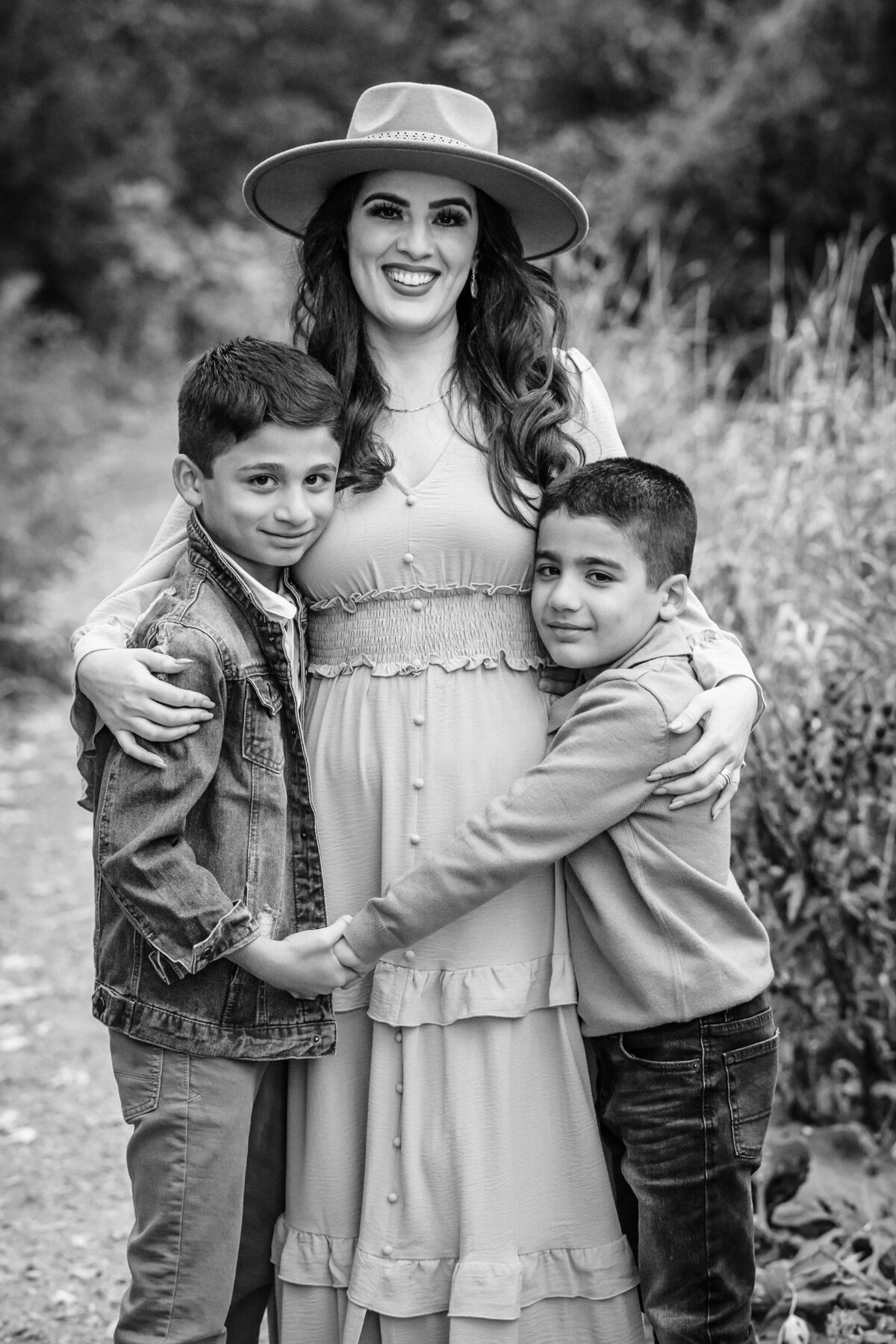 A black-and-white photo of a mother embracing her two young sons, one on each side, while they smile lovingly at the camera. The mother is dressed in a long, elegant dress with a wide-brimmed hat.
