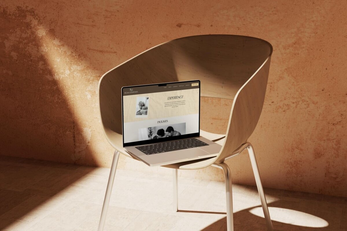 Laptop set on a wooden chair in warm natural light, open to a photography website “Experience” page with a neutral beige design and black-and-white family images.