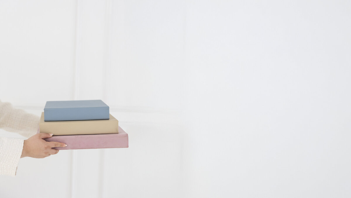 Hands holding a stack of three books in pastel colors: blue, beige, and pink, against a minimalistic white background. Feels serene and organized.