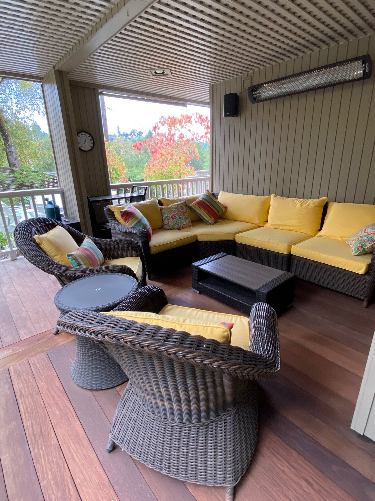 A covered patio with wicker seating, yellow cushions, and wood decking overlooking a landscaped yard.