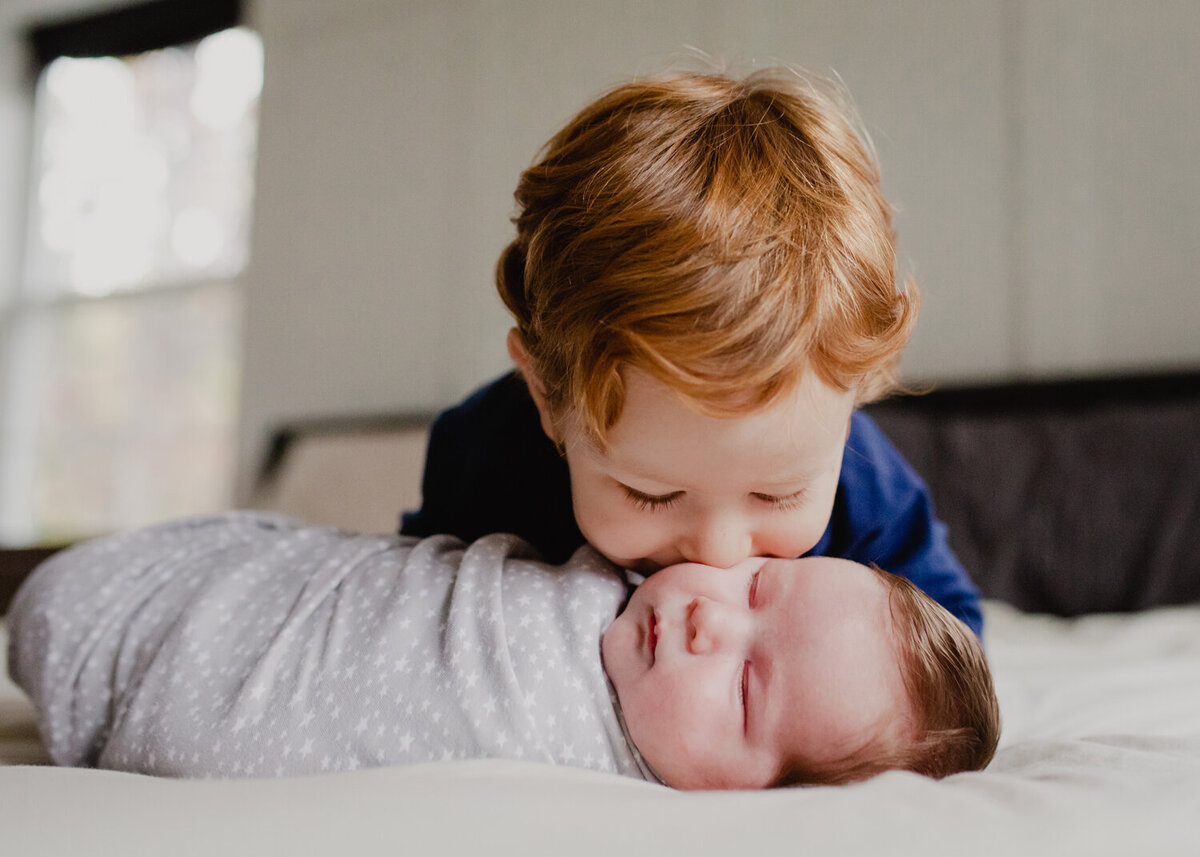 A toddler brother is so excited to meet his new baby brother! He leans over on the bed and kisses his brother while the newborn is swaddled and sleeping.
