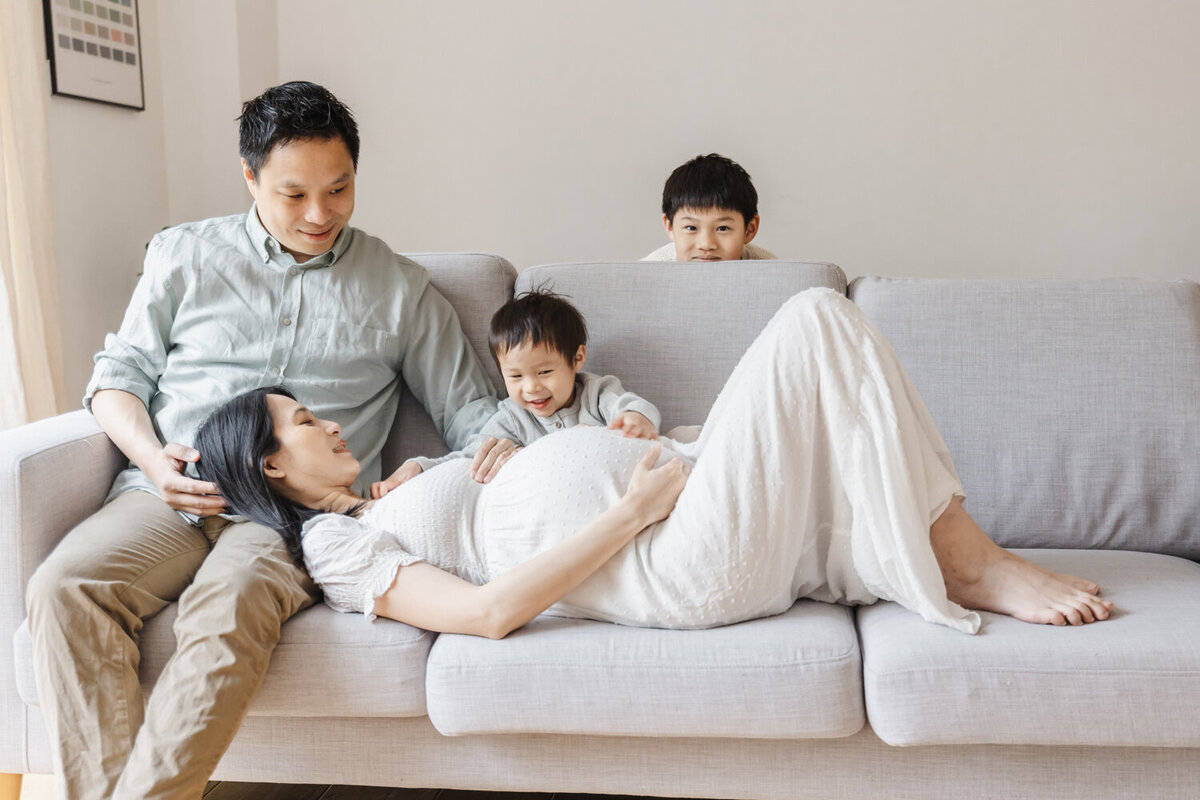 expecting mom laying on couch with her head on her husbands lap. Young son sitting behind her patting her belly. Older son is peeking out from behind the couch.