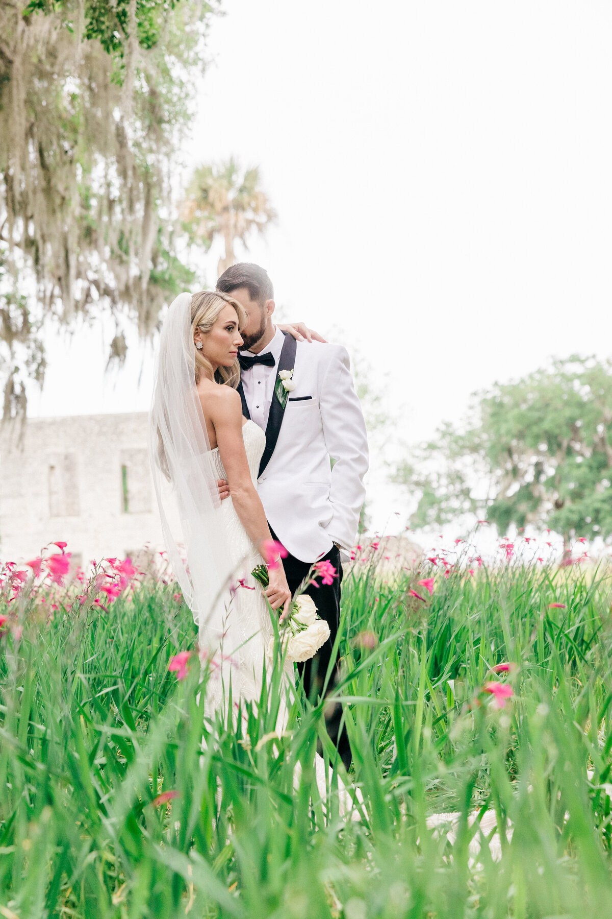 Bride and groom in white-tie attire share a quiet embrace amid wildflowers at Spring Island, South Carolina—captured by luxury wedding photographer Amia Marcell.