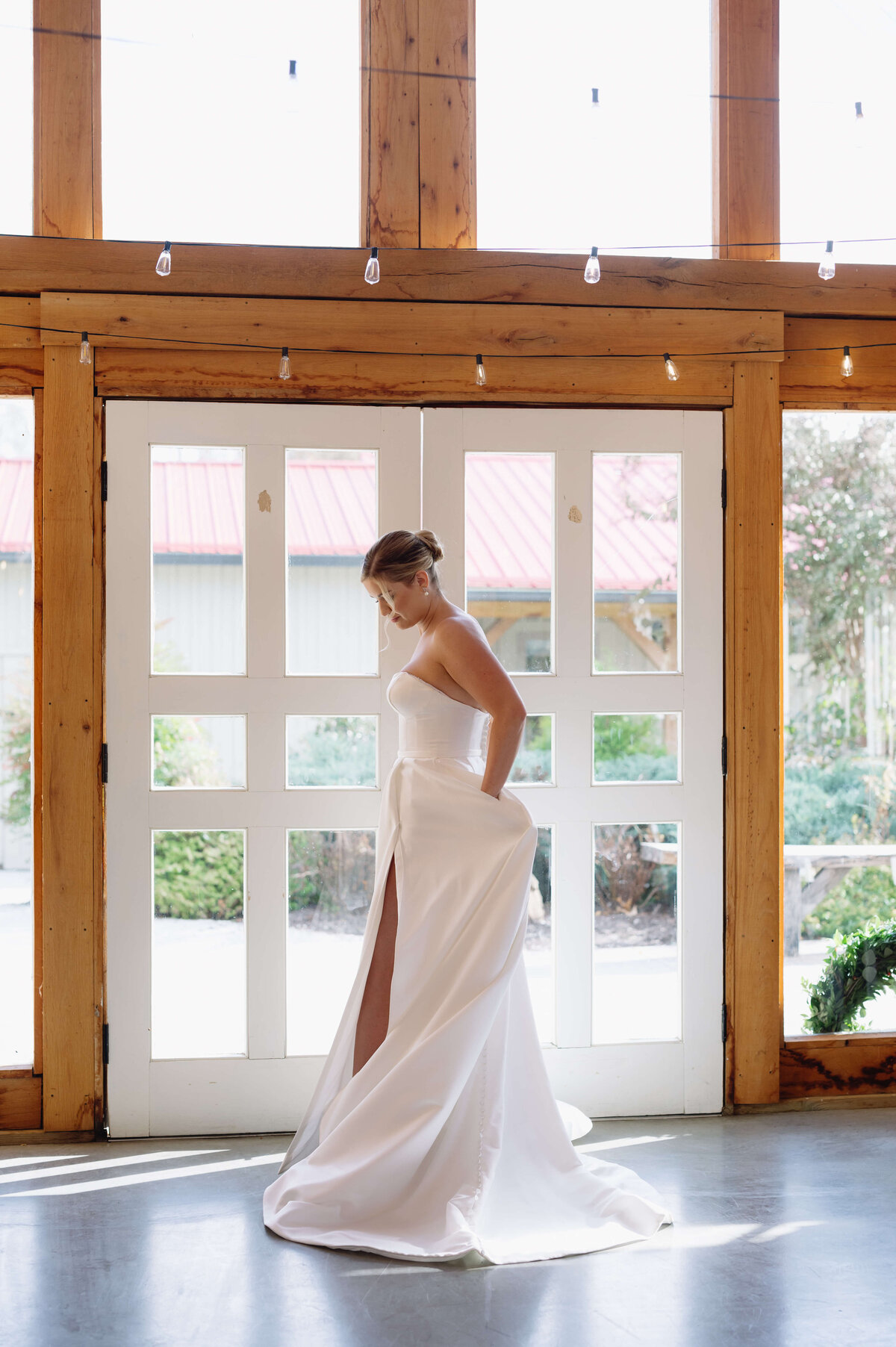 bridal portrait with bride standing in front of a wall filled with windows allowing light to shin in over her as she holds her hands in her gown's pockets with her leg popped out of the slit in her skirt
