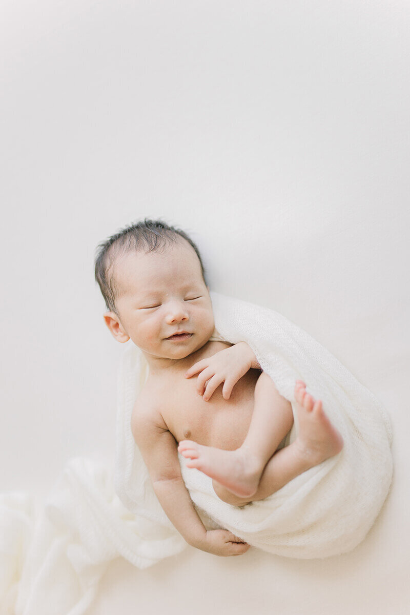 The newborn baby is laying on his back curled up like an egg. He is relaxed and smiling. Shot in Yokohama by Yukika Ishikawa Photography.