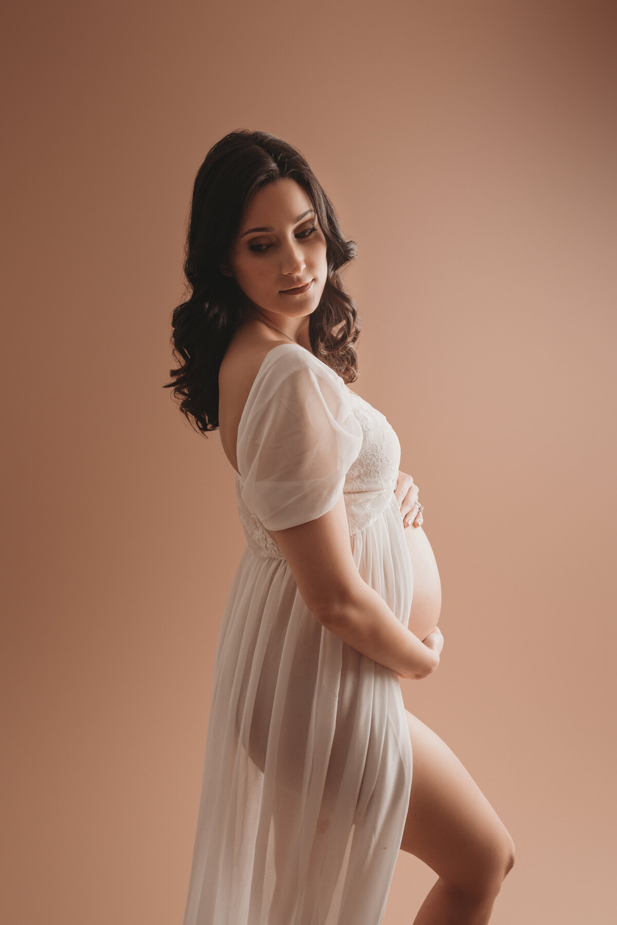 Maternity portrait on tan backdrop of brown haired woman wearing white sheer dress showing her baby bump looking over shoulder