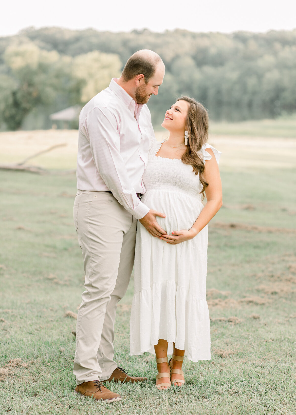 Maternity session in open green grass field. Mom to be is wear long white dress and posing with husband by central mississippi maternity photographer.