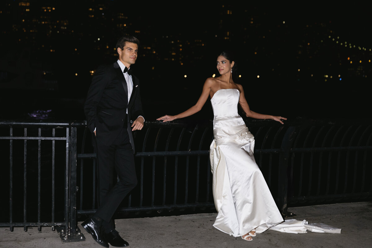 Editorial-style wedding portrait of couple at a waterfront with city lights behind them.