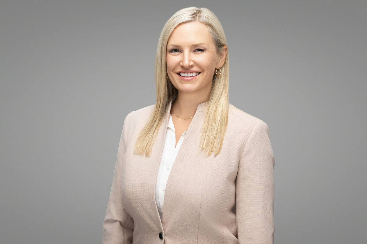 Professional headshot of a smiling blonde woman with straight hair, wearing a light beige blazer over a white blouse, posed against a smooth gray studio backdrop. Captured by Janel Lee Photography in Cincinnati, Ohio, this corporate portrait highlights a confident and approachable expression, ideal for business and personal branding use.