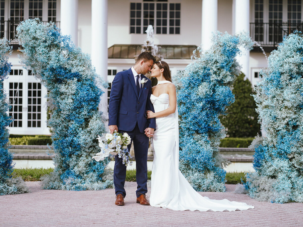 A romantic outdoor wedding moment featuring the bride and groom standing in front of a stunning blue baby’s breath floral structure. The airy and delicate floral installation adds a modern yet ethereal touch, making this a perfect inspiration for couples looking to incorporate bold floral artistry into their special day.