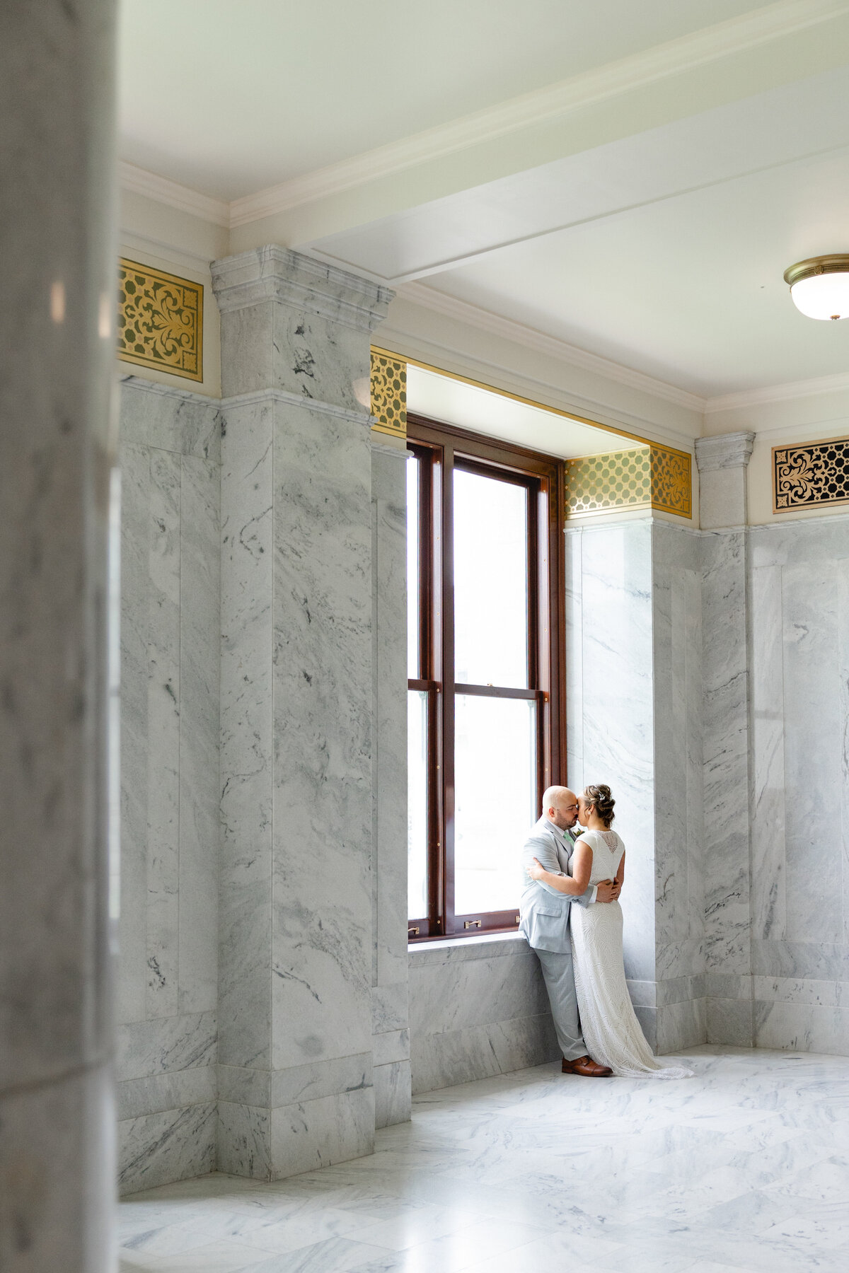 A bride and groom embrace in a grand marble hallway of the Utah State Capitol, surrounded by elegant architecture and soft natural light.