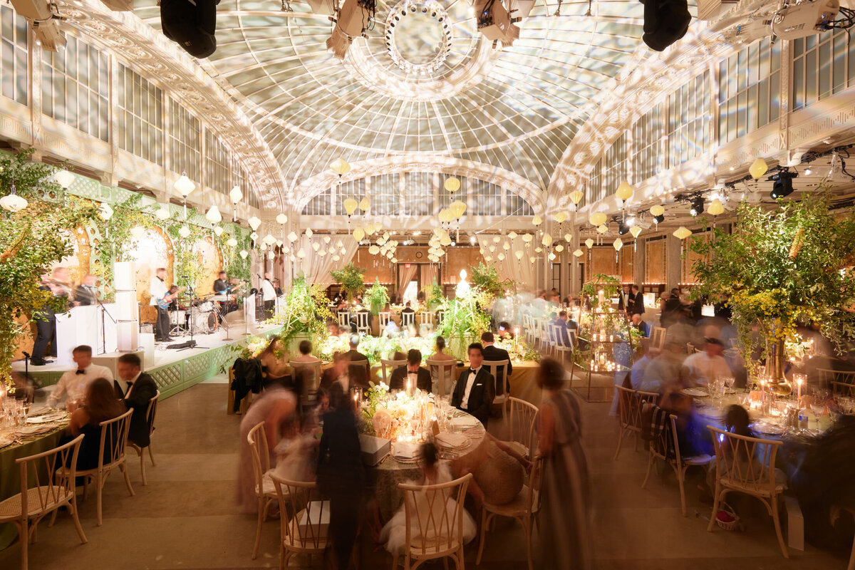 The photo was taken at the New York Public Library in New York City by photographer Shawn Connell. It depicts a large, elegant event in a grand hall with a glass ceiling. The hall is beautifully decorated with abundant greenery and hanging lights, creating a magical ambiance. Round tables are elegantly set for dinner, with guests seated and mingling, enjoying the festive atmosphere. A live band performs on stage, adding to the lively vibe of the event. Aaron Navak Films also captured the event as the Videographers, showcasing their expertise in documenting special moments.