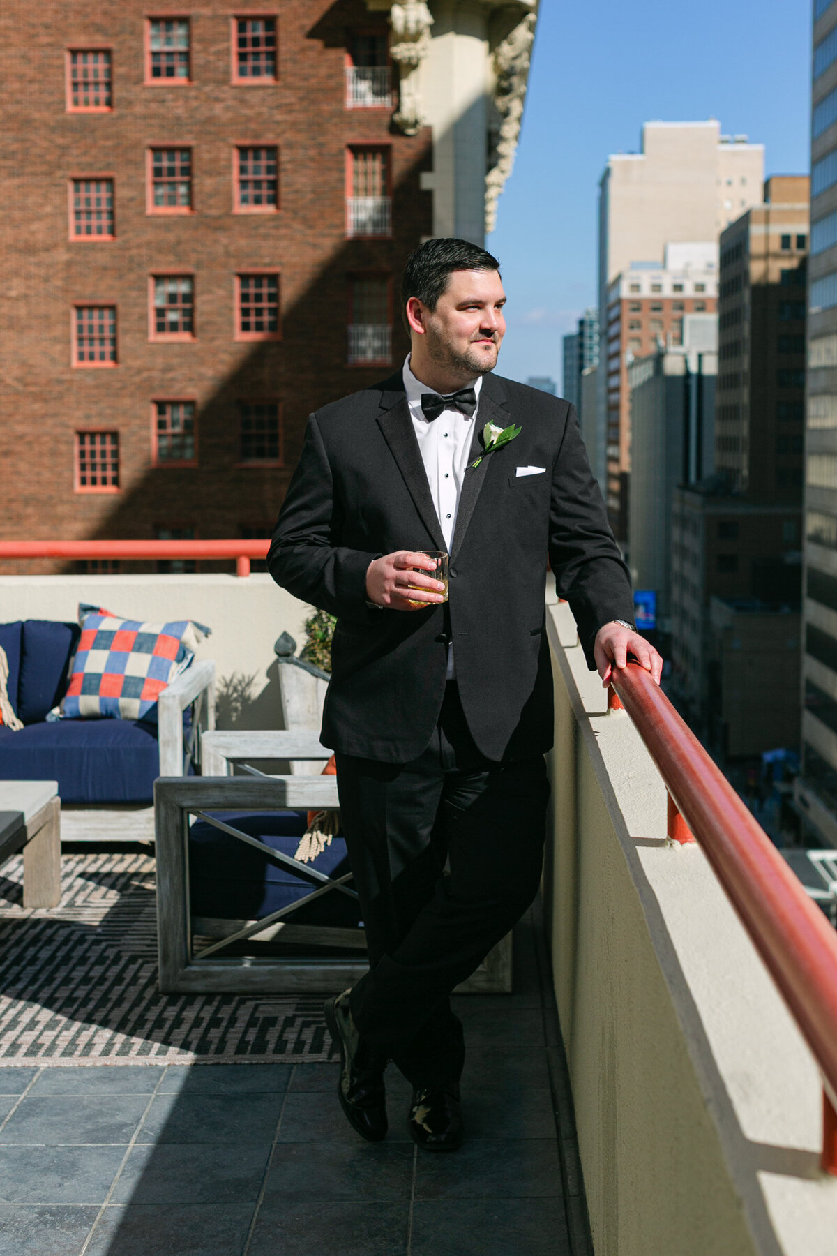 groom standing on a balcony with a whiskey glass in hand at The Adolphus in Dallas, captured in a stylish and elegant wedding portrait.