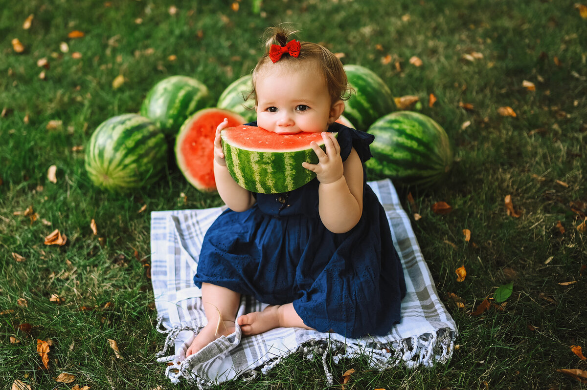 Toddler sits on a blanket on the grass while she eats a slice of watermelon.