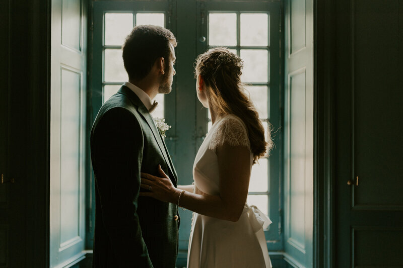 Mariés se faisant face à contre jour se tenant dans l'encadrure d'une fenêtre immortalisés par laura, photographe de mariage en vendée.