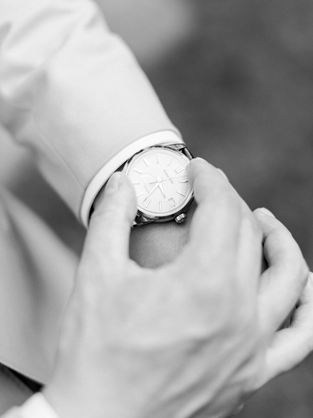 Black and white image of groom adjusting a sleek wristwatch on their left wrist, wearing a light-colored jacket.