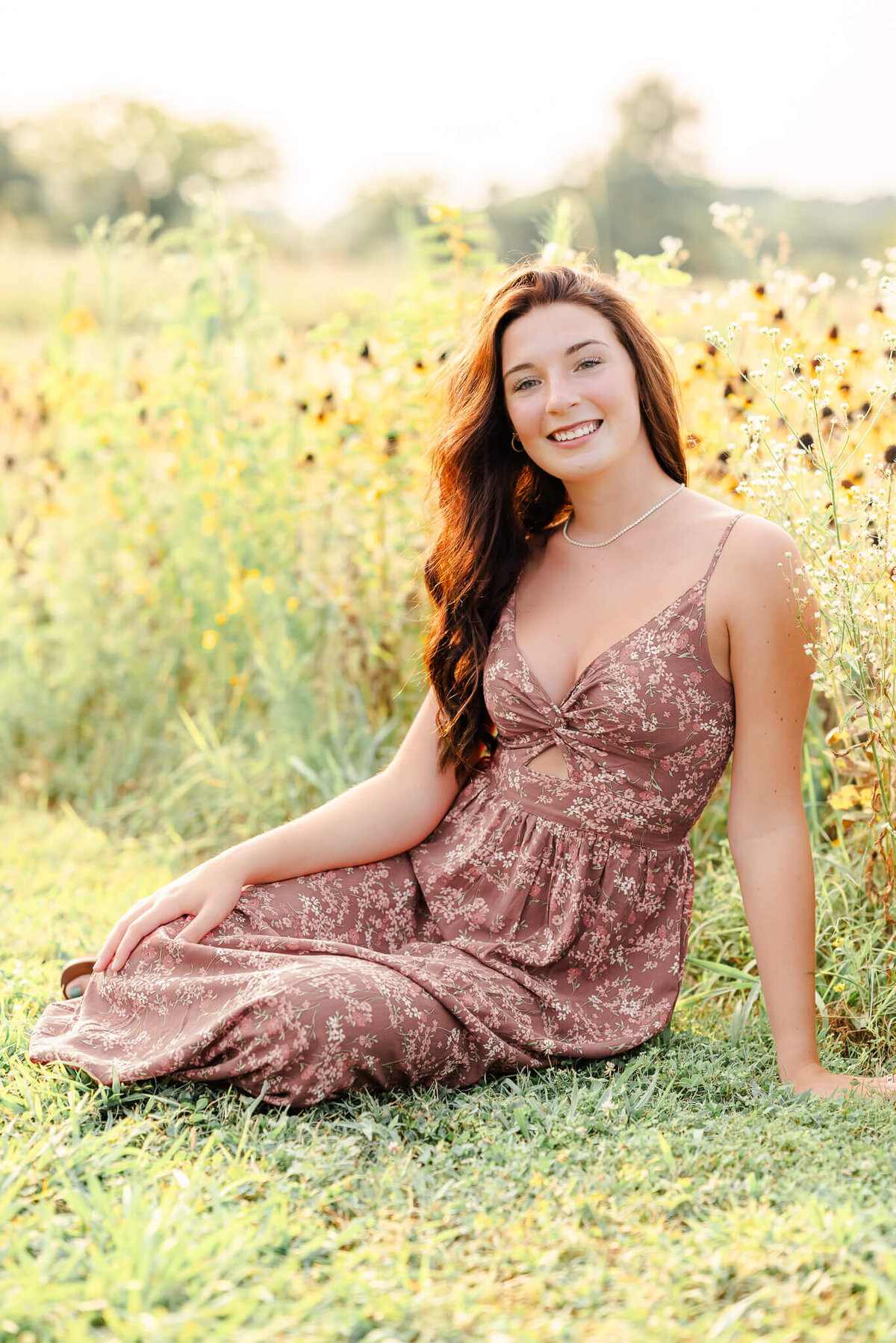 A high school senior, wearing a floral pink dress, sits in front of some yellow flowers during her Chesapeake senior session.